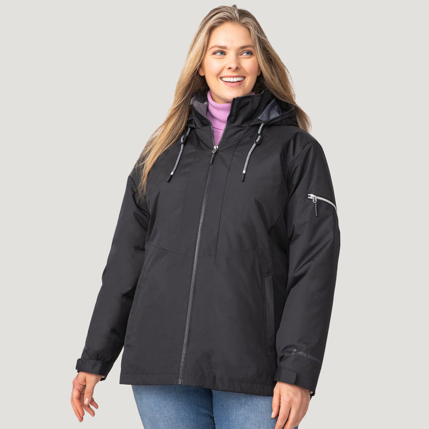 A woman wearing the Clearance Women's Plus Size Back of Bell 3-in-1 Systems Jacket in black, with a hood, paired with light blue jeans, stands and smiles against a plain light gray background.