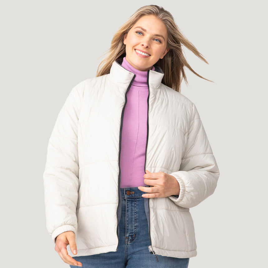 A woman with long blonde hair is wearing a light purple turtleneck, blue jeans, and the Women's Plus Size Back of Bell 3-in-1 Systems Jacket by Clearance—a white sustainable puffer—smiling and glancing to the side against a plain light background.
