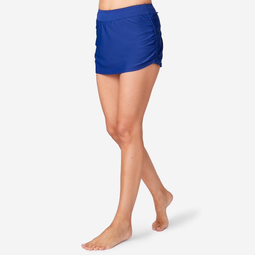 A woman models the Free Country Women's Side Shirred Swim Skirt with adjustable side ties, standing barefoot with one leg forward against a plain light background.