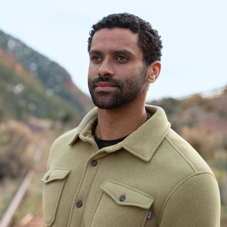 Man wearing a tan button up coat standing outdoors with mountains in the background