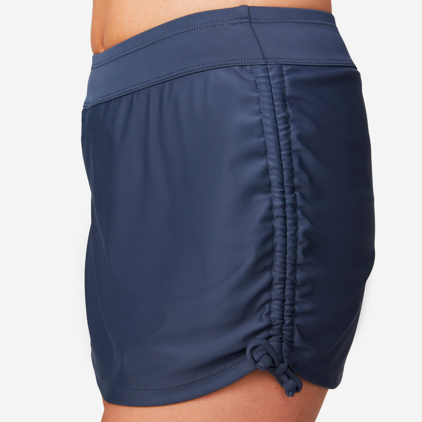 A close-up side view shows a woman wearing the Free Country Women's Side Shirred Swim Skirt, featuring navy blue color, adjustable side ties, a built-in brief, and ruched detail against a light background that highlights its stylish design.