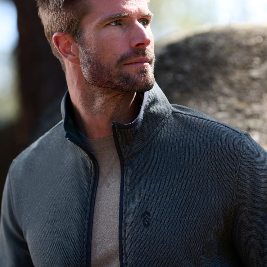 A man with short hair and a beard stands outdoors in front of a large rock, looking to his left while wearing the Men’s Cliffside Pique Fleece Jacket by Free Country in gray over a beige shirt. The jacket’s lightweight warmth makes it perfect for cool outdoor settings.