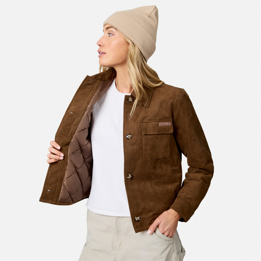 A woman wearing a brown suede-like button-front jacket with a quilted brown lining, a beige beanie, a white T-shirt, and light beige pants, shown against a plain light gray studio background.