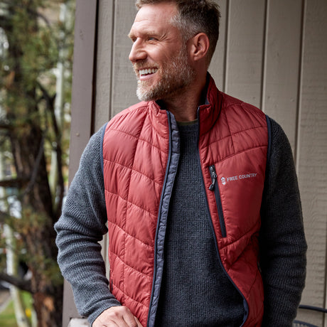 Man wearing the Free Country Men's FreeCycle® Stimson Puffer Vest in blue, featuring quilted design, REPREVE® poly fill, and a vertical zip, layered over a grey long-sleeve shirt.