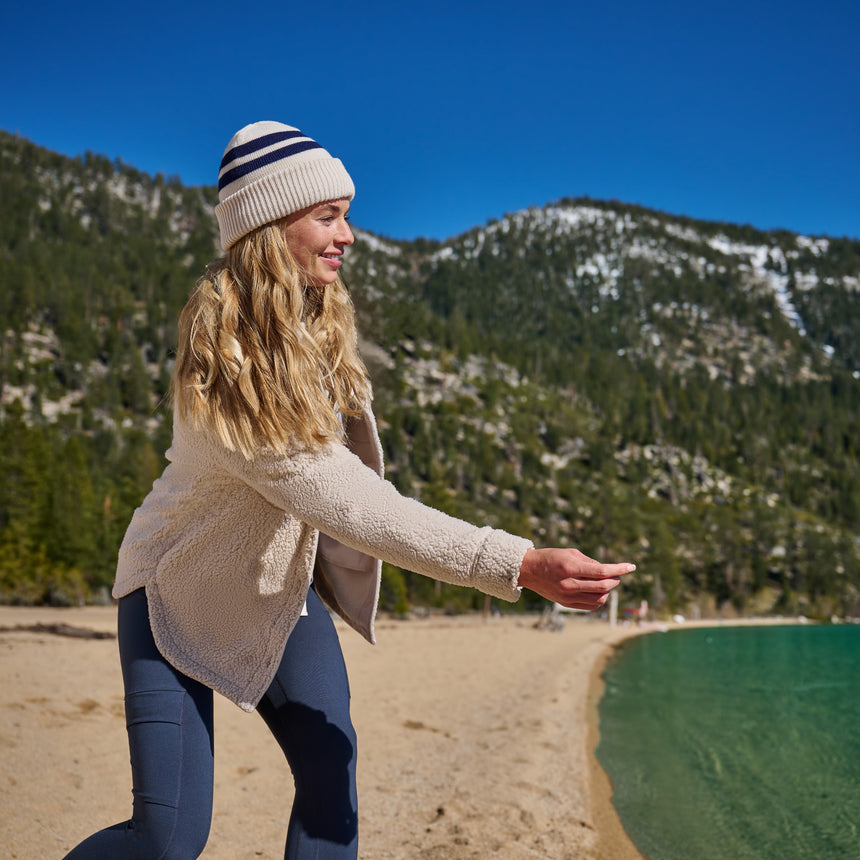 A woman wearing a cream sherpa fleece jacket, navy leggings, and a cream beanie with navy stripes stands on a sandy beach with mountains and a lake in the background.