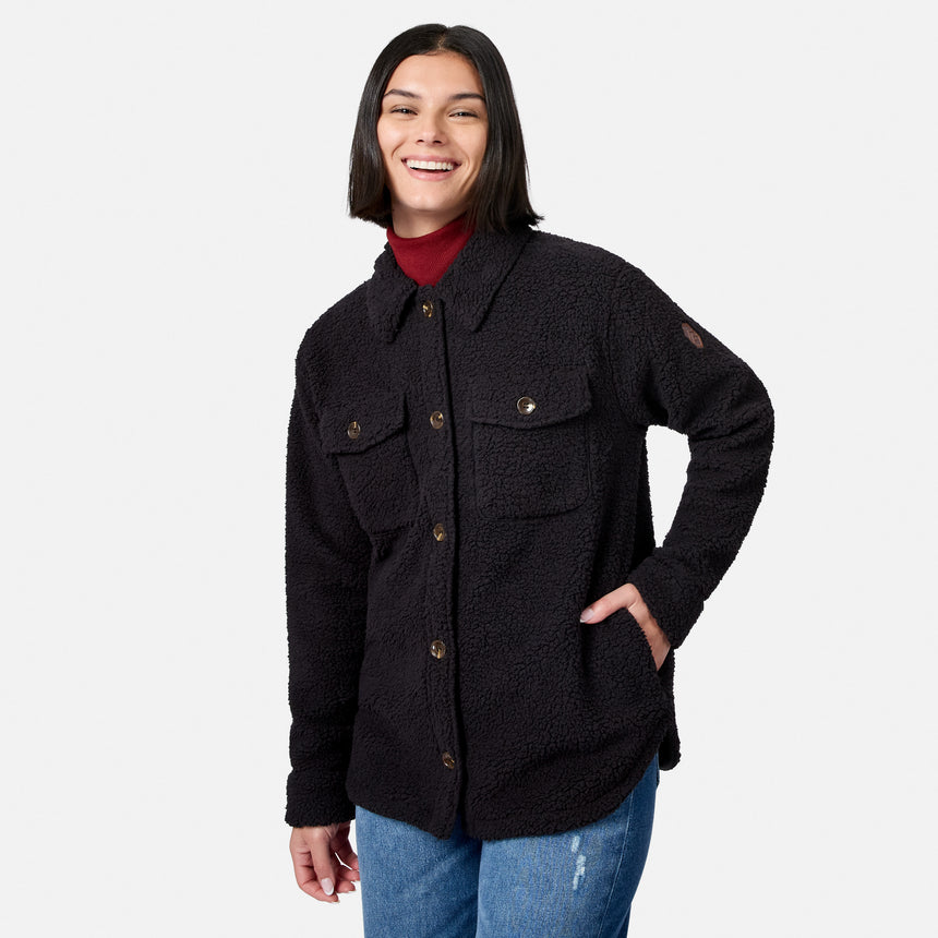 A smiling person with straight dark hair wears the Free Country Women’s Sherpa Shacket in black—crafted from super-soft Sherpa—in an oversized fit over a red turtleneck and blue jeans, posing with one hand in their pocket against a plain light background.