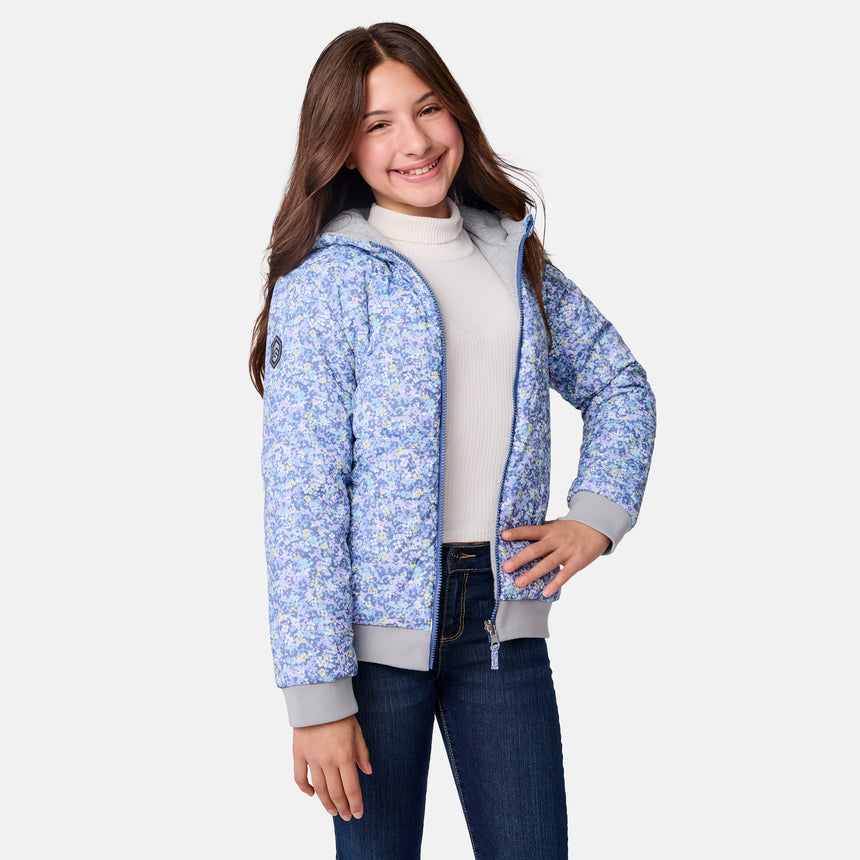 A smiling girl with long brown hair stands against a plain white background, wearing a white turtleneck, dark jeans, and the Clearance Girls' Poofie Reversible Puffer Jacket in a blue floral print. One hand rests on her hip.