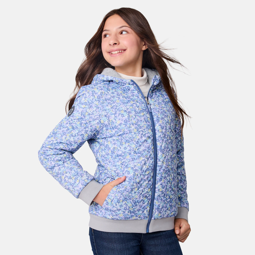 A smiling girl stands with one hand in her pocket against a plain white background, wearing the Clearance Girls' Poofie Reversible Puffer Jacket—a light blue floral zip-up made from recycled polyester with gray cuffs—paired with blue jeans.