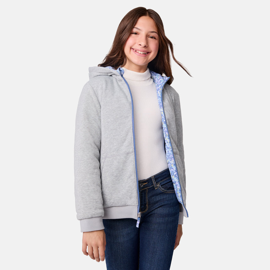 A smiling girl with long brown hair wears the Girls' Poofie Reversible Puffer Jacket by Clearance, styled over a white turtleneck and blue jeans, standing against a plain light background.
