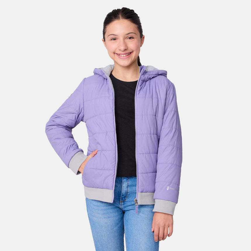 A smiling girl models the Clearance Girls' Poofie Reversible Puffer Jacket in light purple, worn over a black shirt and blue jeans, standing with one hand on her hip against a plain white background.