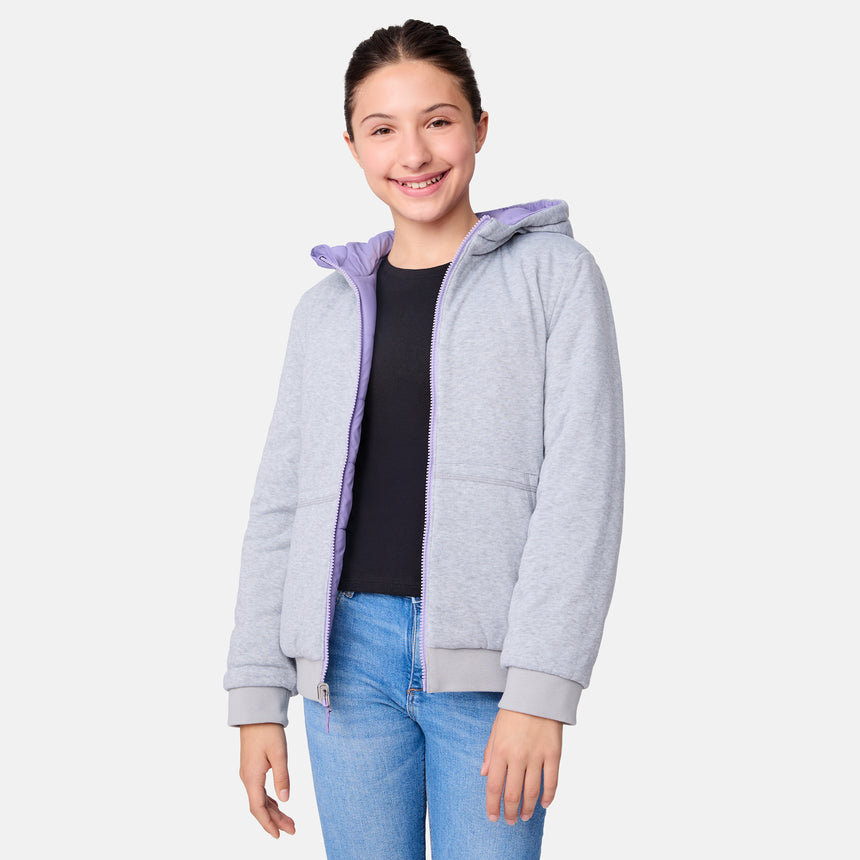 A smiling young person stands against a plain white background, wearing the Clearance Girls' Poofie Reversible Puffer Jacket in light gray over a black shirt and blue jeans.