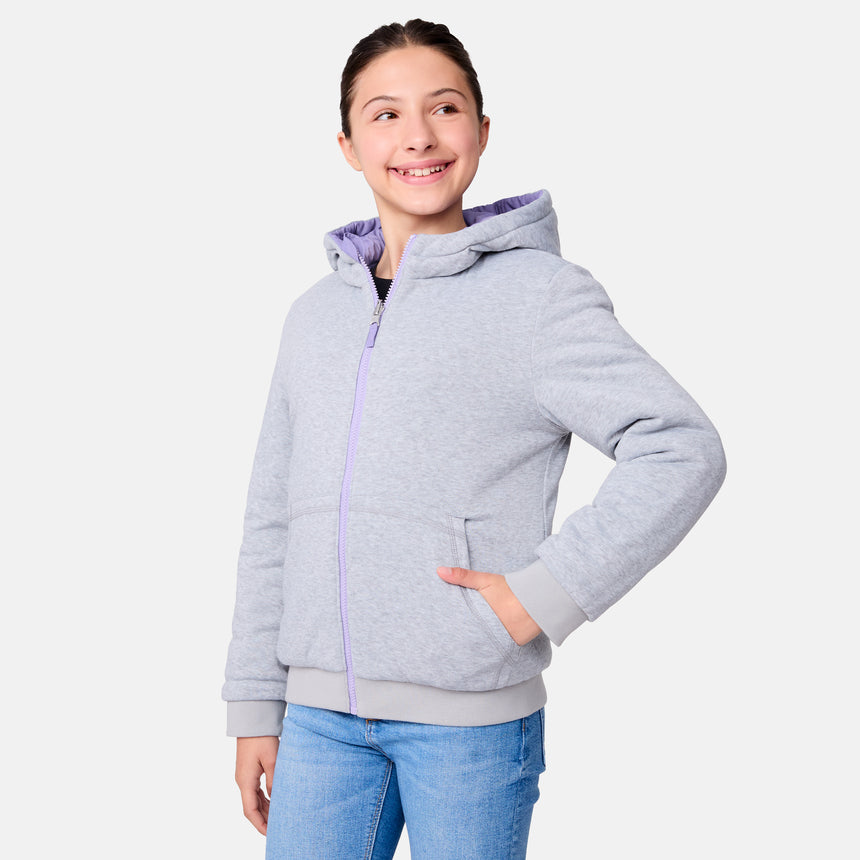 A young girl wearing the Clearance Girls' Poofie Reversible Puffer Jacket in light gray with a lavender lining and blue jeans smiles while looking to her left, posing with one hand in her pocket against a plain white background.