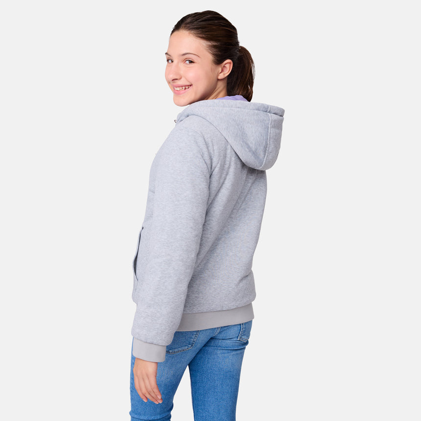 A young woman in a gray hooded sweatshirt and blue jeans stands smiling, turning partially toward the camera against a plain white background, highlighting the versatility of the Girls' Poofie Reversible Puffer Jacket by Clearance.