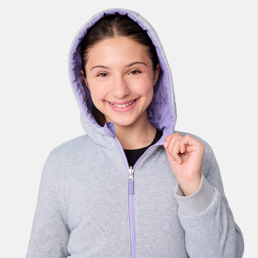 A young person with long dark hair smiles while wearing the Girls' Poofie Reversible Puffer Jacket by Clearance in light gray with a lilac lining, holding the zipper. The background is plain white.