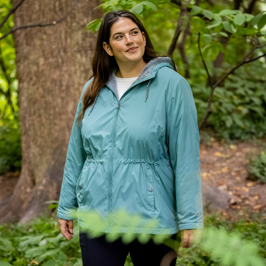 Woman wearing Free Country Women's Cascade Windshear Jacket in light blue with a full front zipper opening, soft lining, and water resistant and wind resistant recycled polyester stretch performance fabric. Best women’s jacket for windy weather.