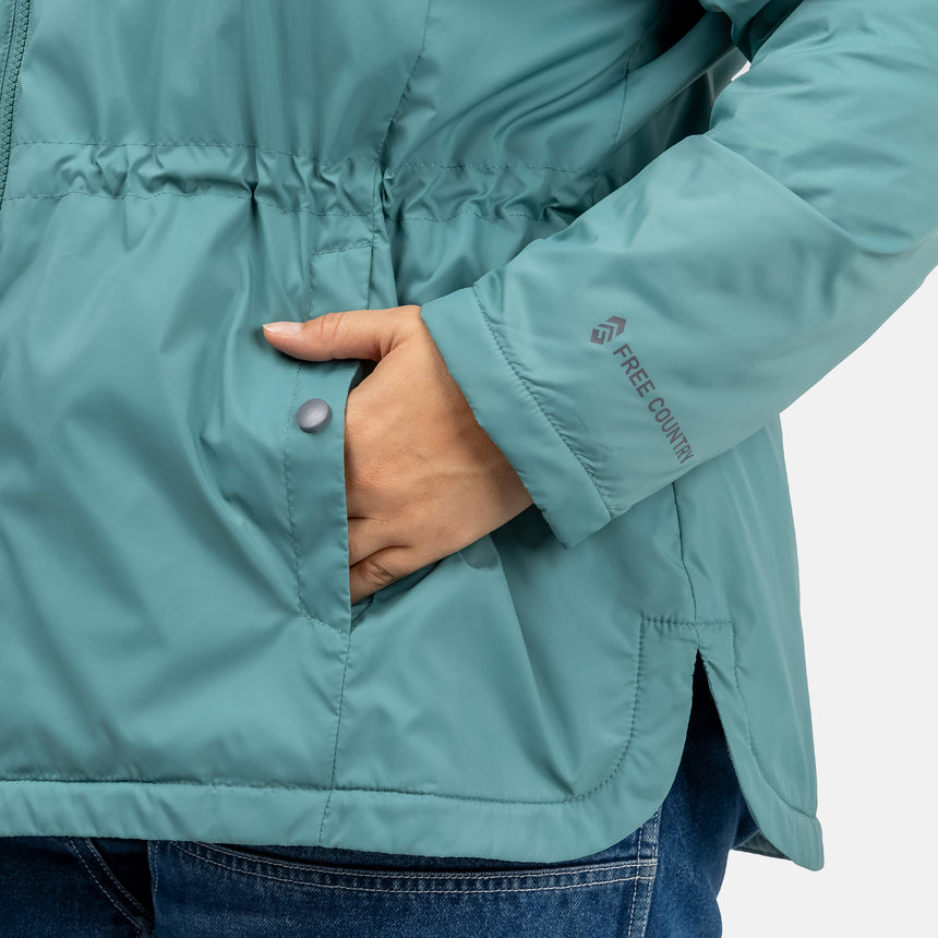 Woman wearing Free Country Women's Cascade Windshear Jacket in light blue with a full front zipper opening, soft lining, and water resistant and wind resistant recycled polyester stretch performance fabric, light grey background. Best women’s jacket for windy weather.