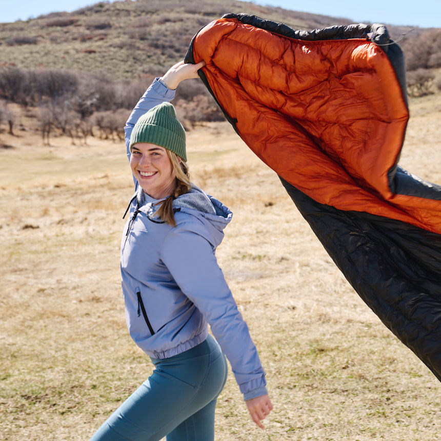 Woman in blue Free Country windbreaker holding an orange and black sleeping bag in a field