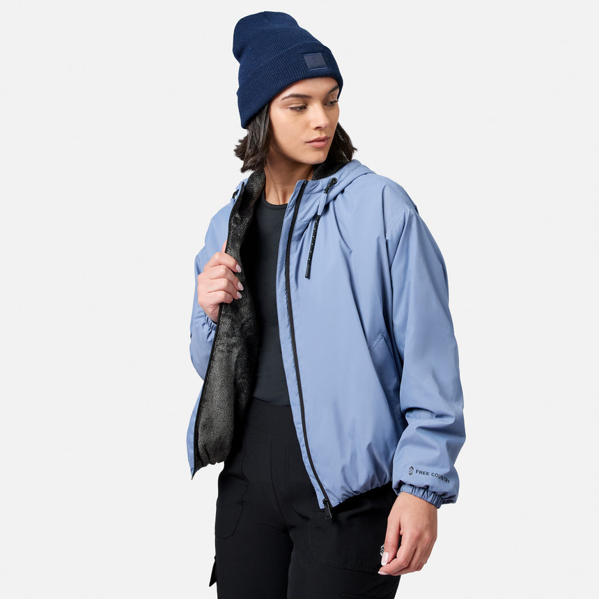 Model wears the Free Country Women’s Bomber Windshear Jacket in light blue with black lining, paired with a blue beanie and black pants. She looks left, holding the jacket open against a plain white background.