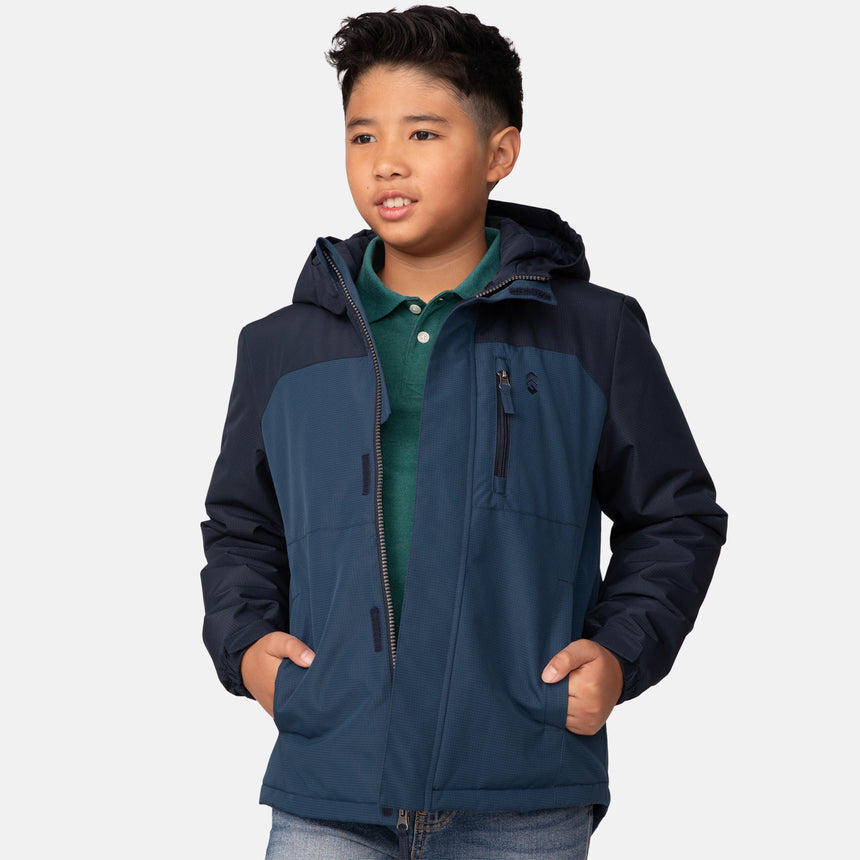 A boy stands with his hands in his pockets, looking slightly to the side and smiling, dressed in a Clearance Boys' Mount Avon Midweight Jacket in blue layered over a green polo shirt.