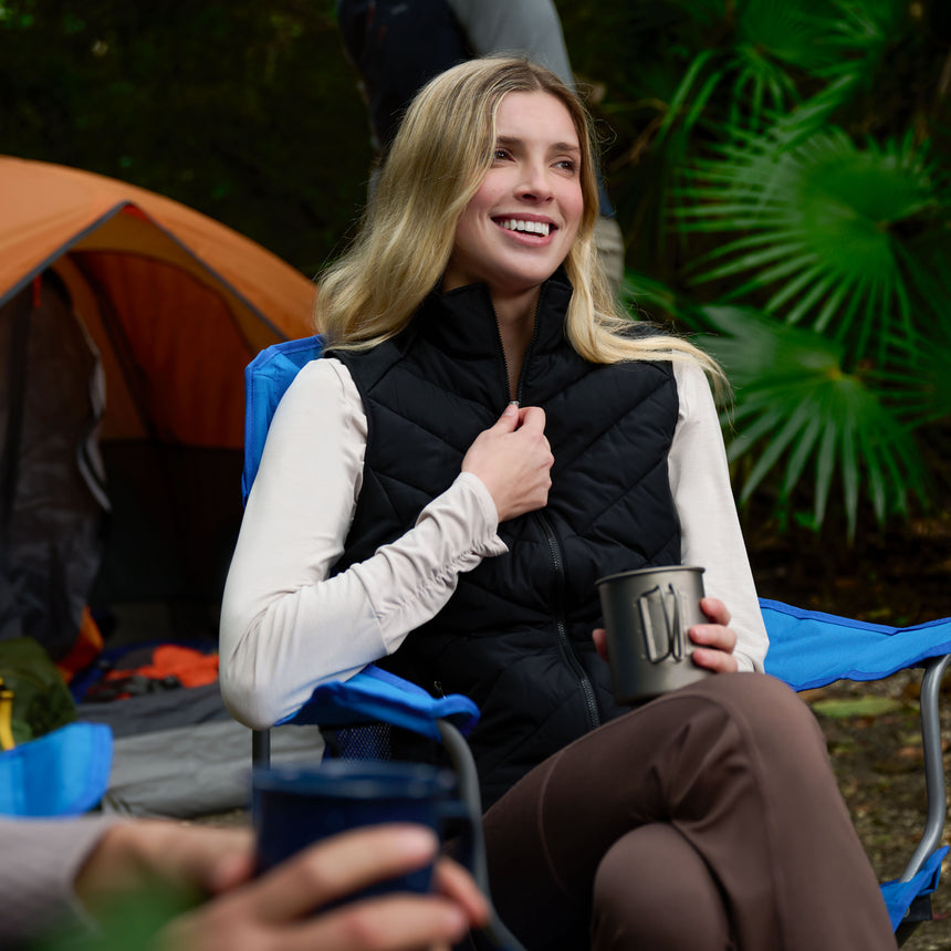 Woman sitting outdoors with a camping chair, holding a mug, surrounded by greenery and a tent.