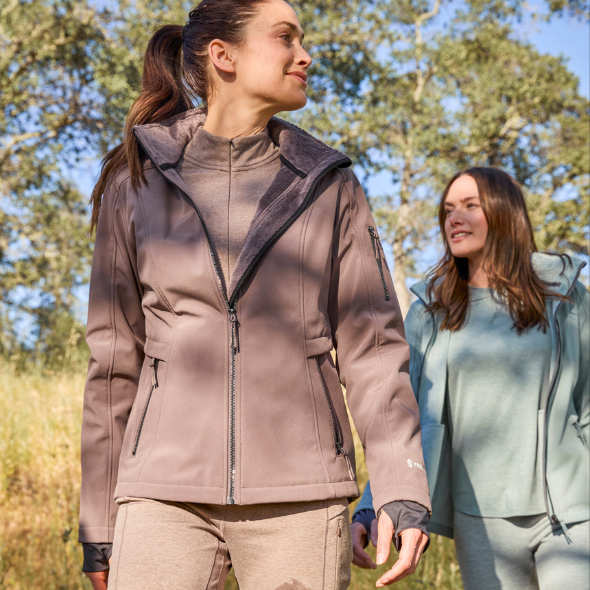 Woman wearing Free Country Women's Aeris II Super Softshell® Jacket in light brown with a full front zipper opening, soft lining, detachable hood, and water resistant and wind resistant stretch performance fabric.