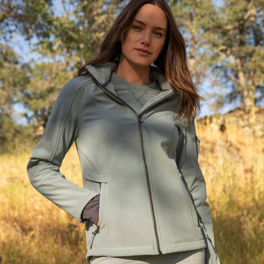 Woman wearing Free Country Women's Aeris II Super Softshell® Jacket in light blue with a full front zipper opening, soft lining, detachable hood, and water resistant and wind resistant stretch performance fabric.