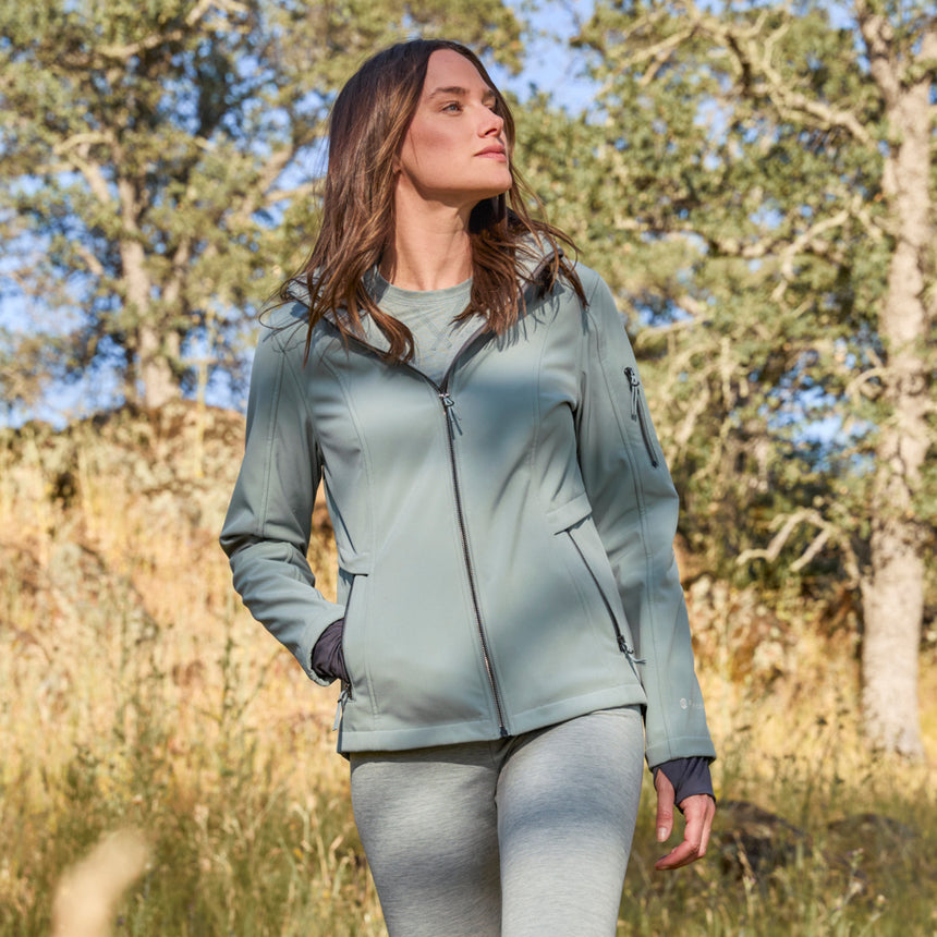 Woman wearing Free Country Women's Aeris II Super Softshell® Jacket in light blue with a full front zipper opening, soft lining, detachable hood, and water resistant and wind resistant stretch performance fabric.