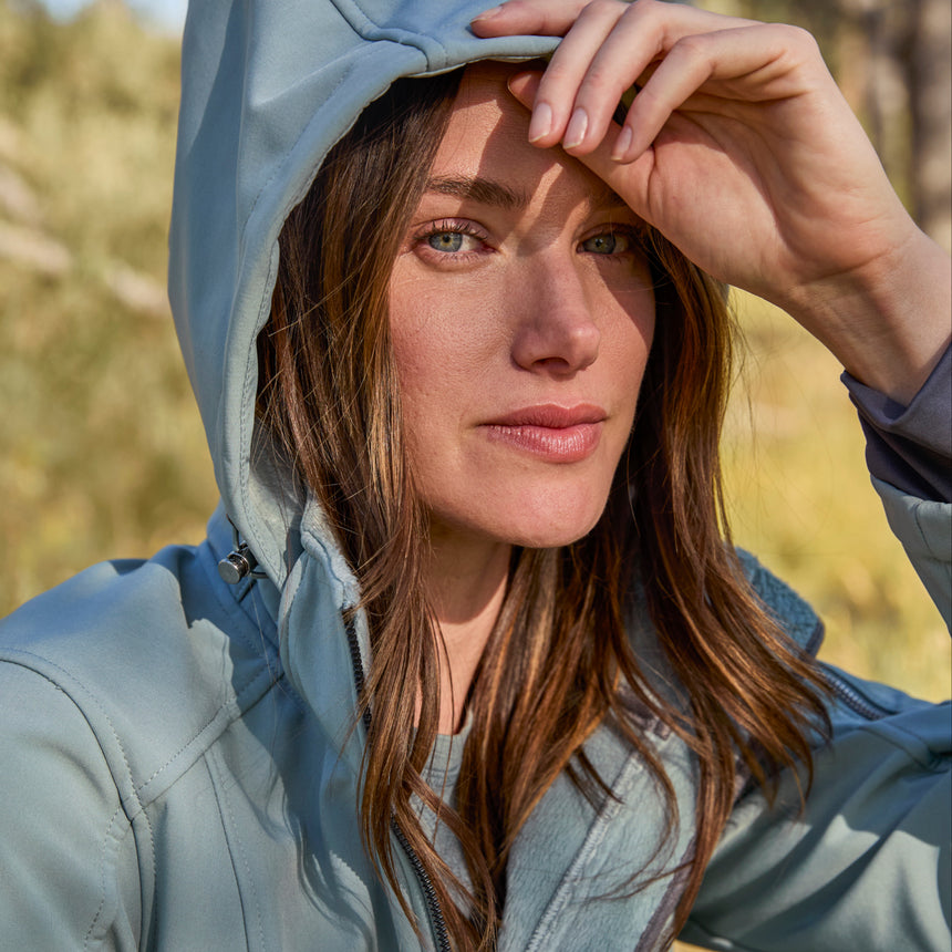 Woman wearing Free Country Women's Aeris II Super Softshell® Jacket in light blue with a full front zipper opening, soft lining, detachable hood, and water resistant and wind resistant stretch performance fabric.