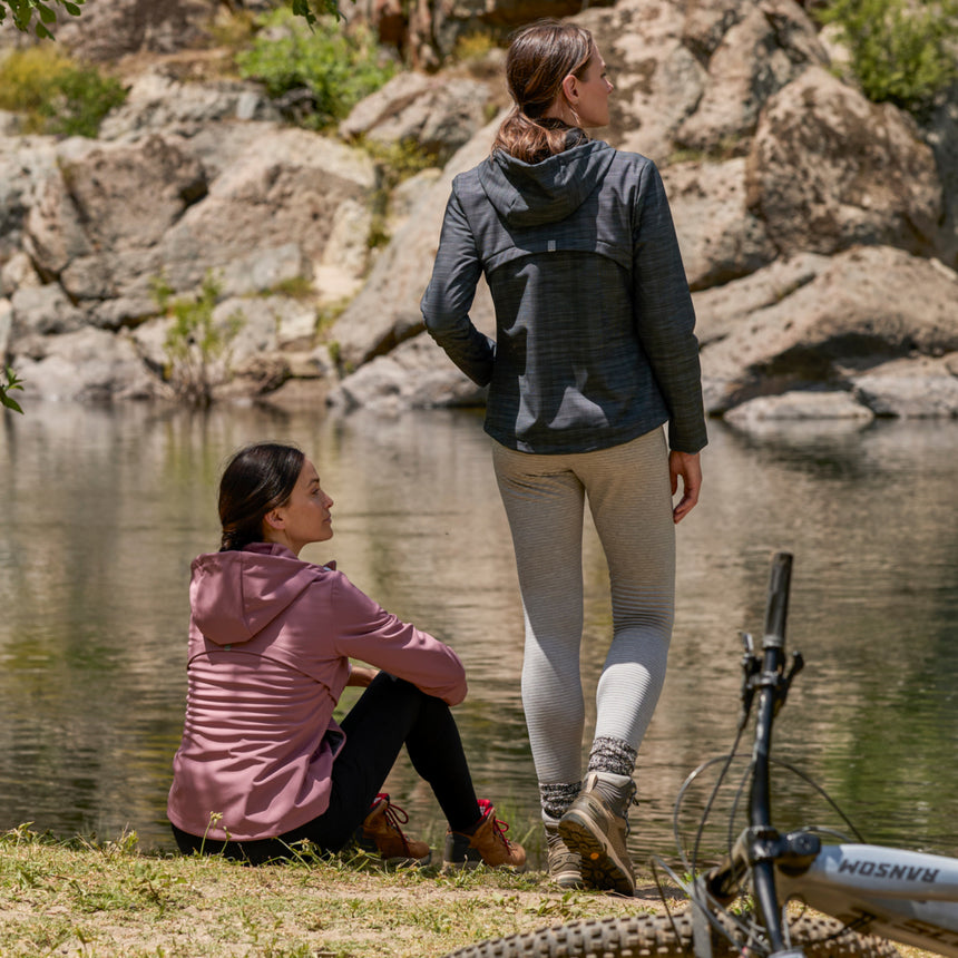 Woman wearing Free Country Women's Alps Super Softshell® Jacket in pink with a full front zipper opening, soft lining, attached hood with adjustable drawcords, and water resistant and wind resistant stretch performance fabric, light grey background. Perfect women’s  jacket for fall weather.