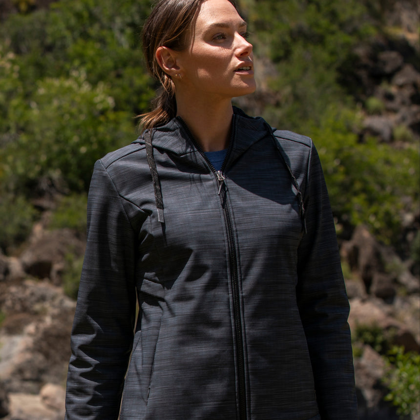 Woman wearing Free Country Women's Alps Super Softshell® Jacket in textured black with a full front zipper opening, soft lining, attached hood with adjustable drawcords, and water resistant and wind resistant stretch performance fabric, light grey background. Perfect women’s  jacket for fall weather.
