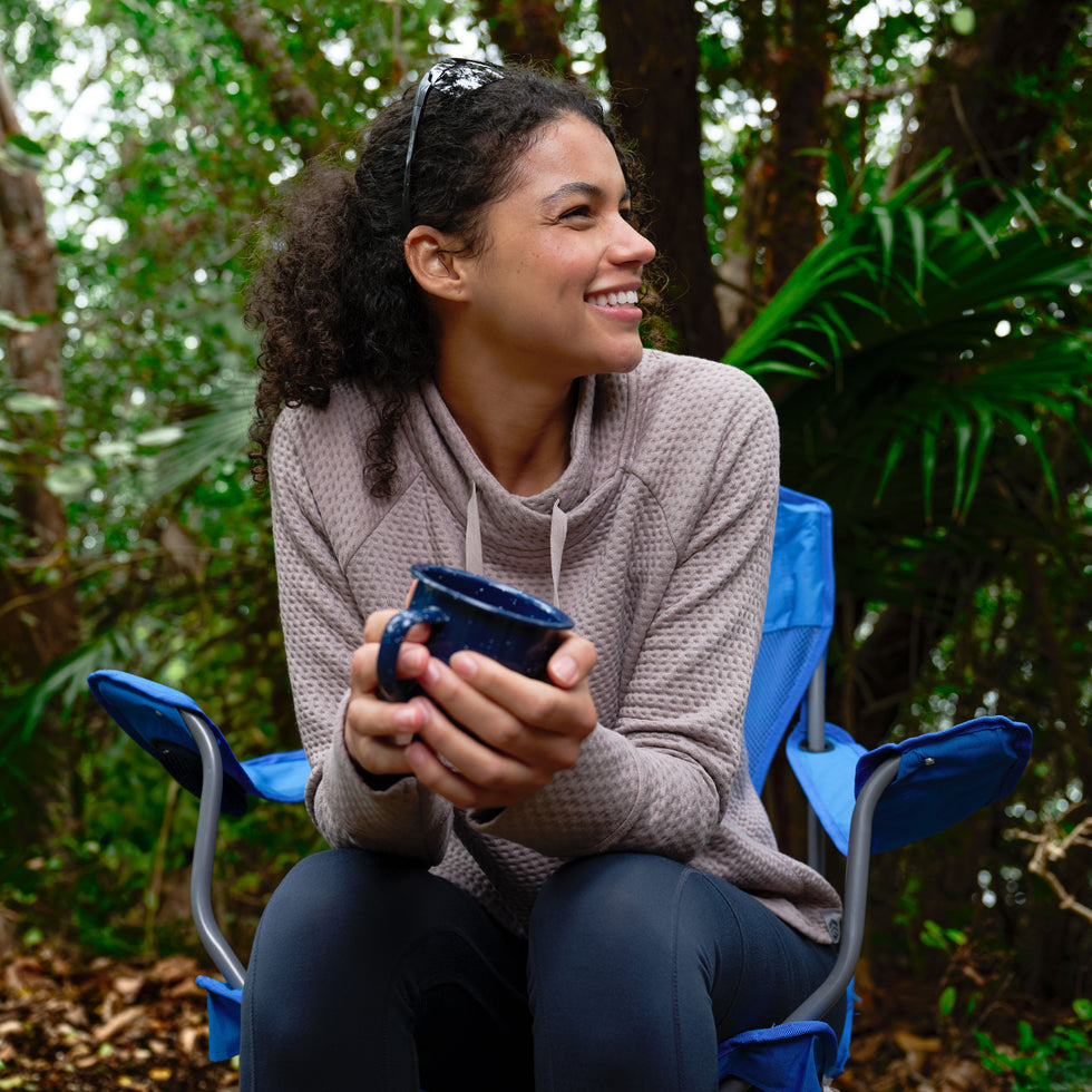 A woman sits outdoors on a blue camping chair, smiling and holding a navy mug. She wears the Free Country Women's UltraFill Cowl Neck Top, featuring light polyester fill and seamless quilting, surrounded by green trees.