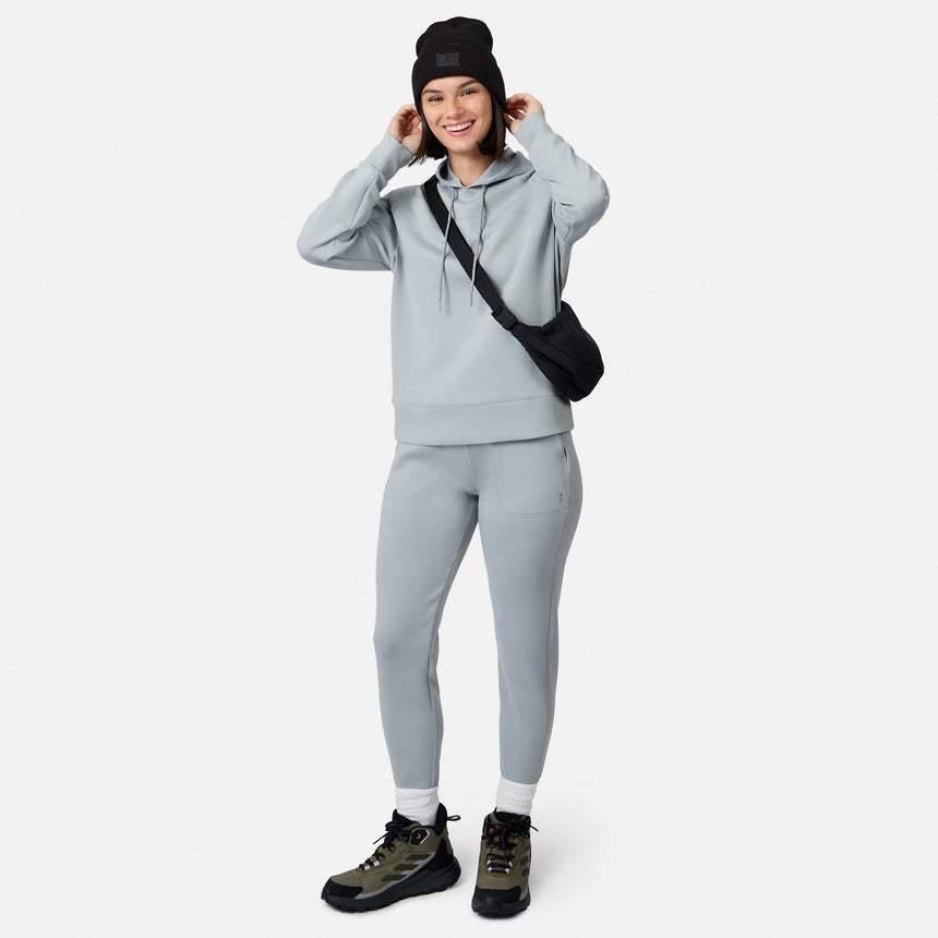 A woman in a light gray Women’s Sueded Lush Pullover Hoodie by Free Country and matching joggers, with a black beanie, crossbody bag, and hiking boots, smiles with hands near her face against a plain white background.
