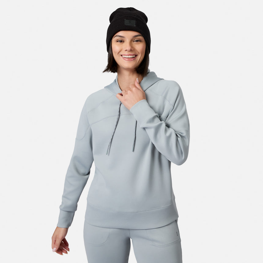 A woman in a Free Country Women’s Sueded Lush Pullover Hoodie and matching pants smiles at the camera, wearing a black beanie and adjusting her hood. The cozy hoodie pops against the plain white background.