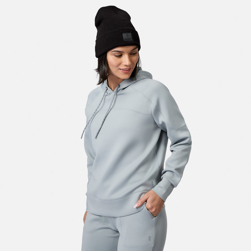 A woman wears the Free Country Women’s Sueded Lush Pullover Hoodie in Lichen, hand in pocket, with a black beanie against a white background, looking down and smiling.