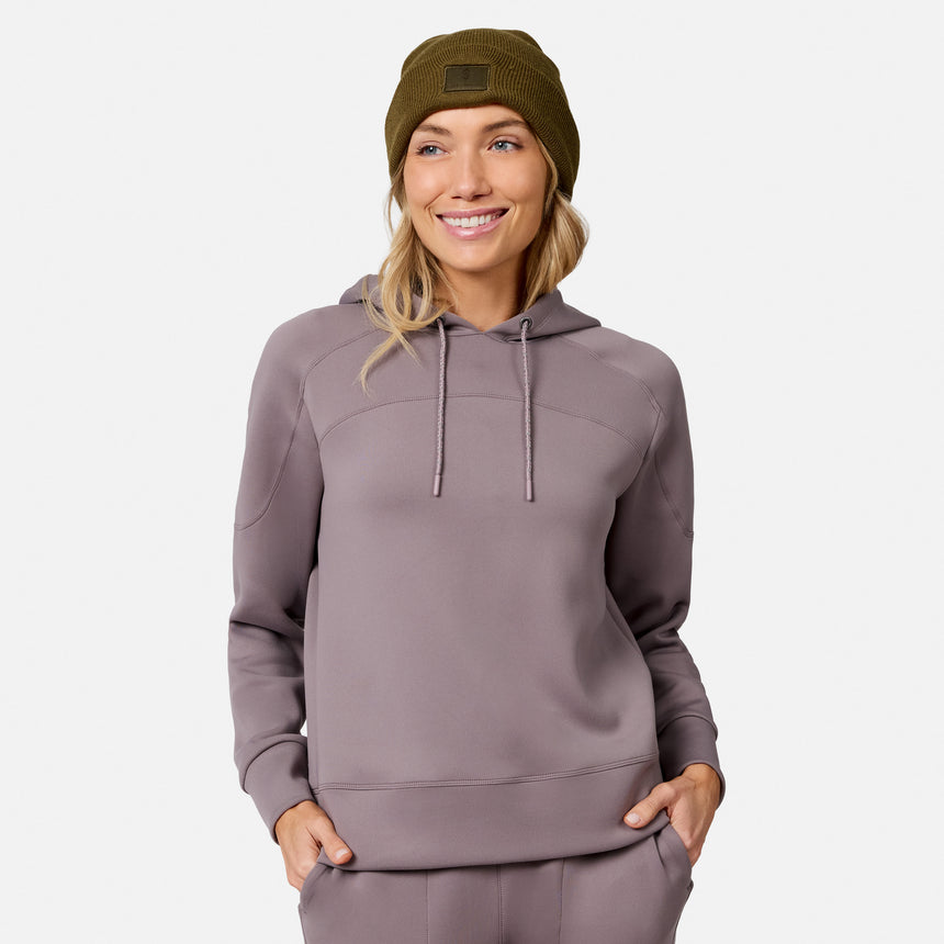 Smiling woman wears the Free Country Women’s Sueded Lush Pullover Hoodie in taupe with matching pants and an olive green beanie, hands in pockets, against a plain white background.