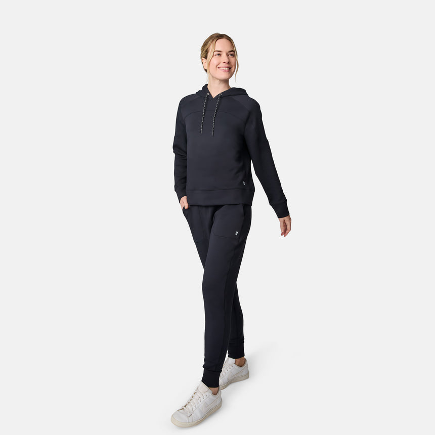 A woman smiles while walking in a matching black Free Country hoodie and Women’s Sueded Lush Jogger, paired with white sneakers, against a plain white background.