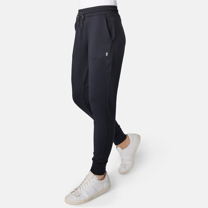 A woman wearing Free Country Women’s Sueded Lush Jogger in black with front pockets and white sneakers, standing against a plain white background.