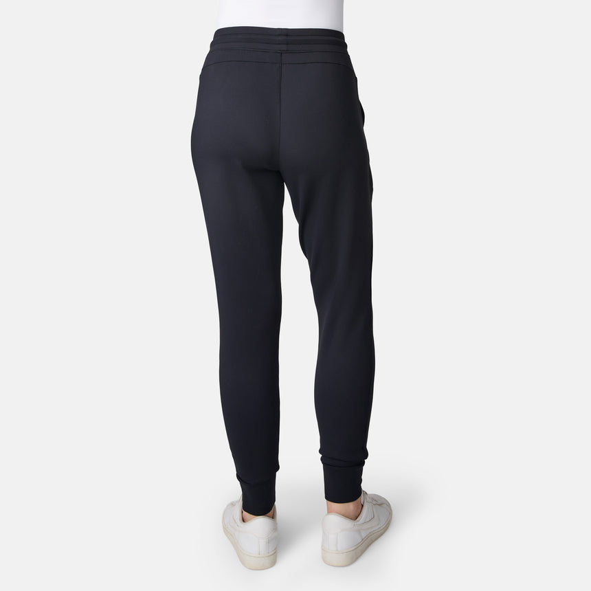 A woman is shown from the back wearing Free Country Women’s Sueded Lush Jogger in black with white sneakers, standing on a plain light background.