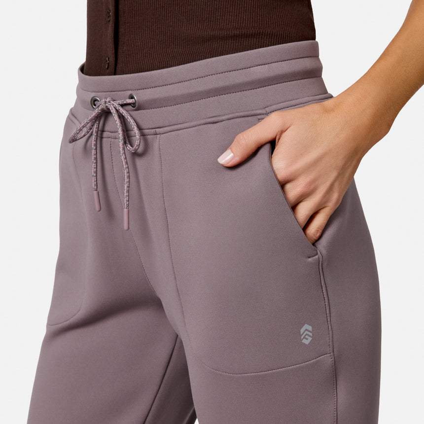 A person wearing Free Country Women’s Sueded Lush Jogger in mauve, featuring 4-way stretch and a drawstring, with their right hand in the pocket. Only the lower torso and right arm are visible against a plain background.