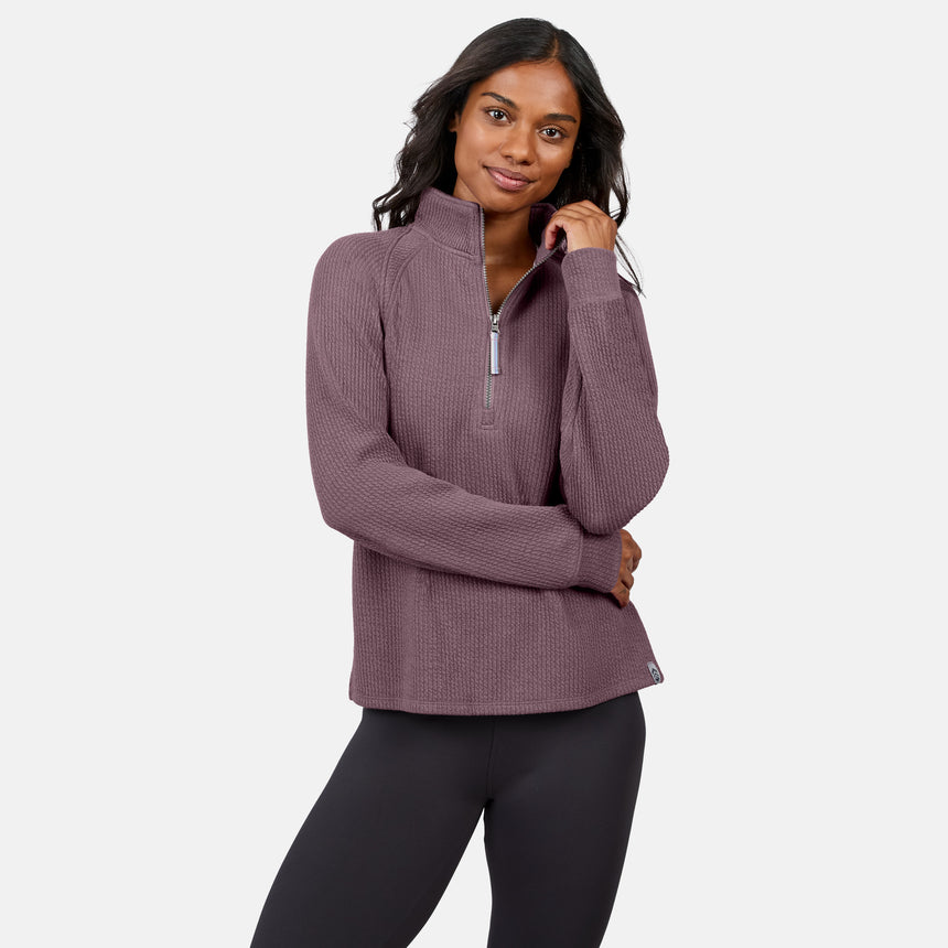 A woman wears the Free Country Women’s Ripple Knit Half Zip Top in mauve with gray leggings, standing against a white background, smiling with one hand in her hair—an ideal pick for casual layering.
