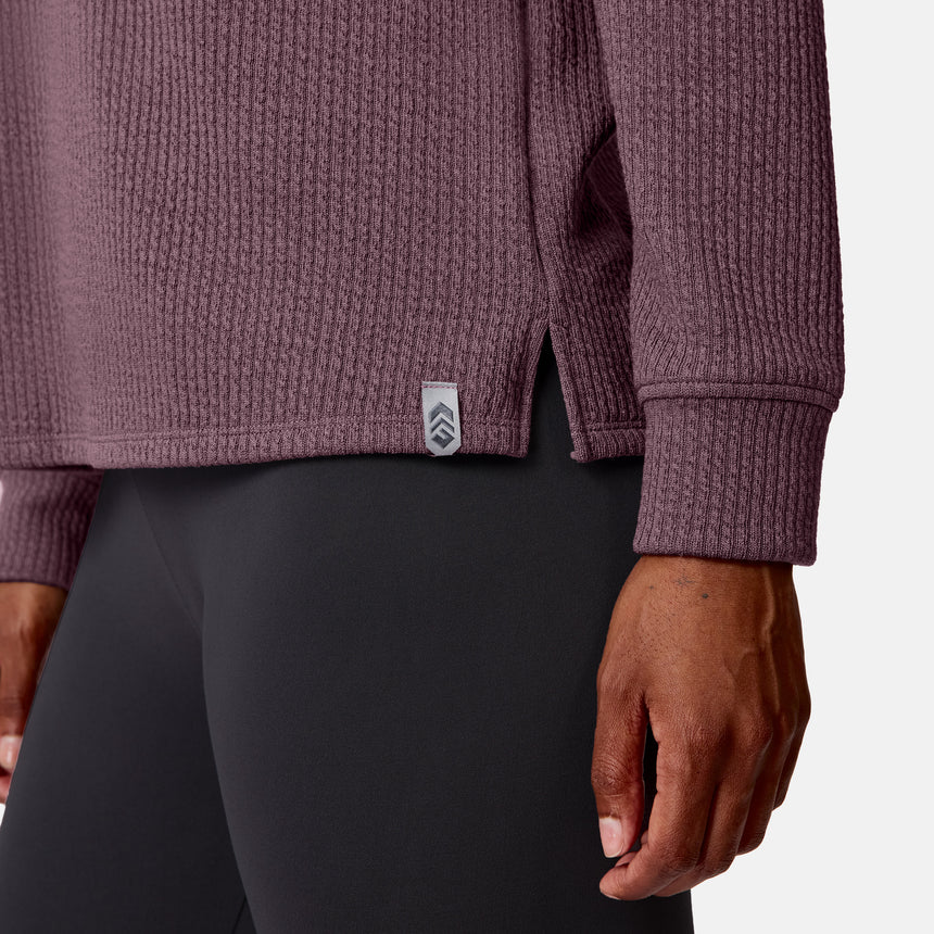 A woman wears the Free Country Women’s Ripple Knit Half Zip Top—a mauve, textured, breathable long-sleeve with a side tag—paired with fitted gray leggings, shown waist down with her right arm by her side.