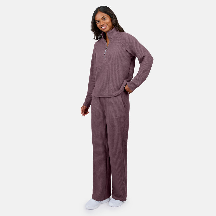 A woman smiles in Free Country’s Women’s Ripple Knit Half Zip Top and matching mauve pants, with her hand in her pocket. She pairs the breathable outfit with white slippers, perfect for layering against a plain white background.