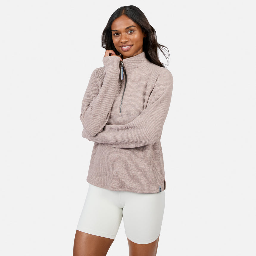 A woman with long dark hair wears a Free Country Women’s Ripple Knit Half Zip Top in Taupewood and white shorts, standing against a plain white background and smiling with one hand touching her chin.