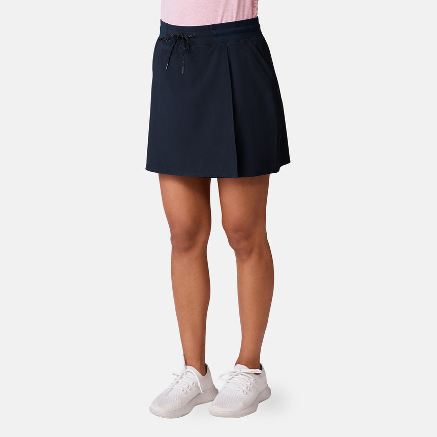 A woman stands against a light background wearing the Free Country Women's Trail to Town Skort in navy blue with a drawstring waist, subtle pleats, moisture-wicking four-way stretch fabric, paired with a light pink top and white sneakers.