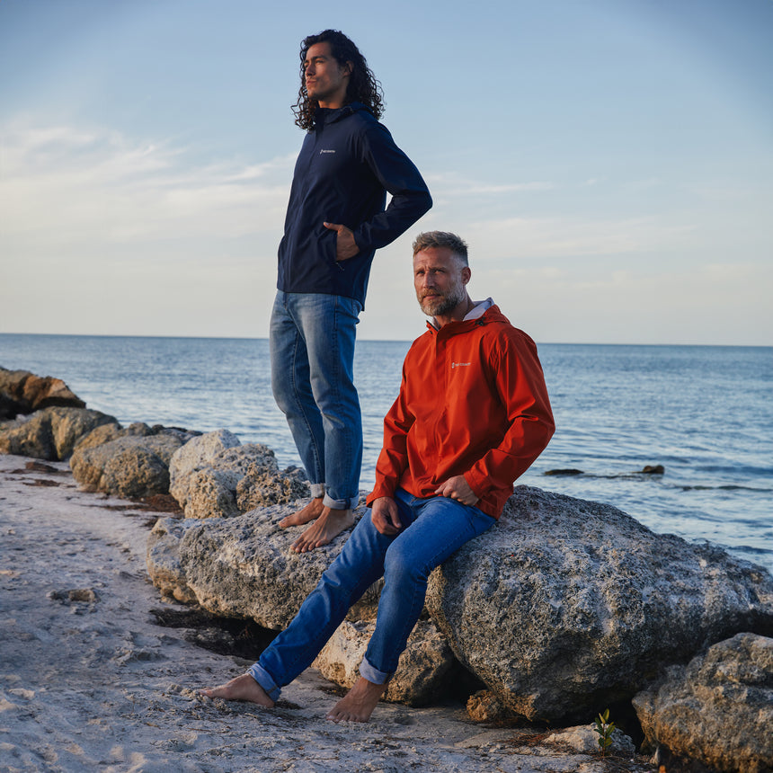 Man wearing Free Country Men's Hydro Lite X2O Spectator Waterproof Jacket in Harris Orange, size M, made from lightweight four way stretch performance fabric featuring sealed seams, elastic cuffs, and an attached hood with a drawcord, styled with blue jeans #color_harris-orange