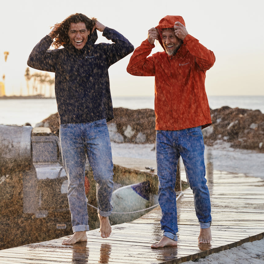 Man wearing Free Country Men's Hydro Lite X2O Spectator Waterproof Jacket in Harris Orange, size M, made from lightweight four way stretch performance fabric featuring sealed seams, elastic cuffs, and an attached hood with a drawcord, styled with blue jeans #color_harris-orange