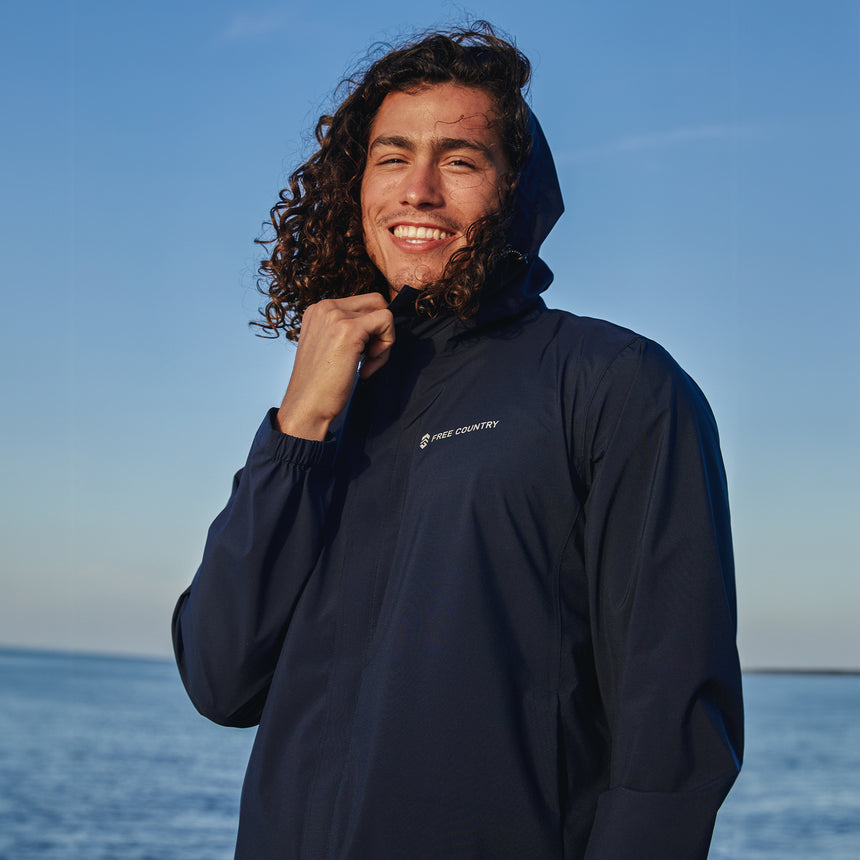 Man wearing Free Country Men's Hydro Lite X2O Spectator Waterproof Jacket in Black, size M, made from lightweight four way stretch performance fabric featuring sealed seams, elastic cuffs, and an attached hood with a drawcord, styled with blue jeans #color_black