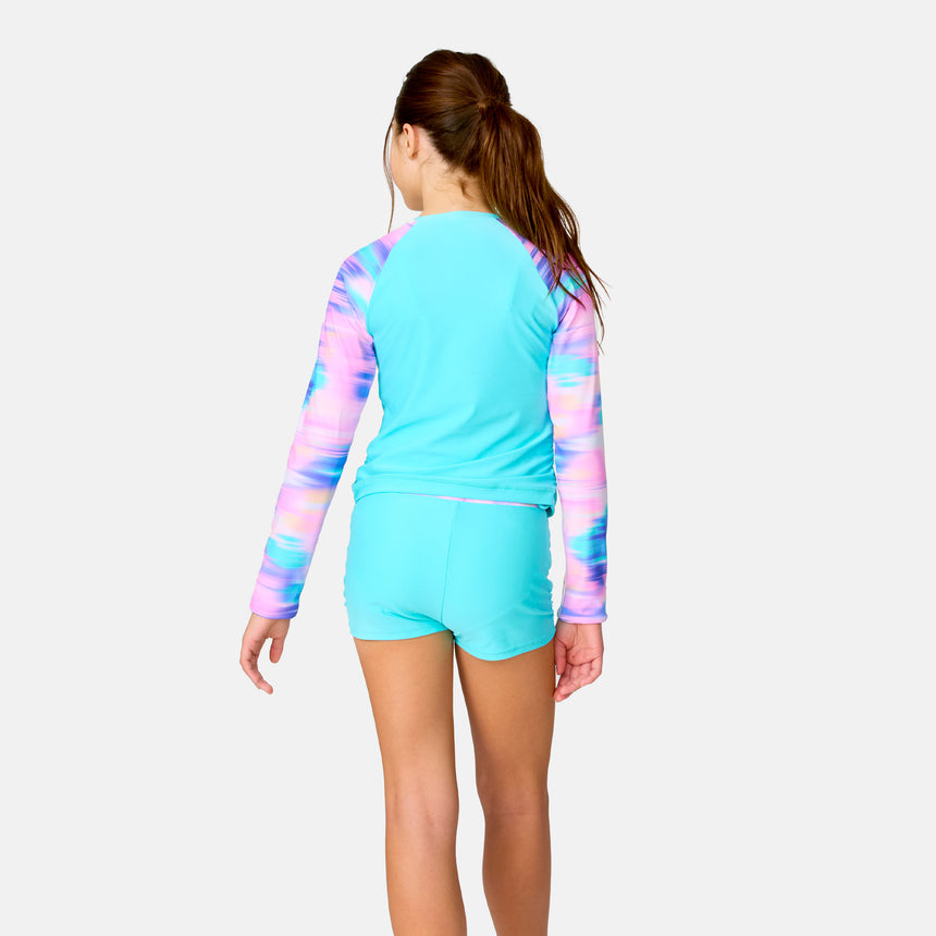 A girl faces away in the Clearance Girls' Dreamy Drift Rash Guard Swim Set, featuring a turquoise UPF 50+ long-sleeve top with pink, purple, and blue tie-dye sleeves and matching quick-dry boy shorts. The background is plain white.