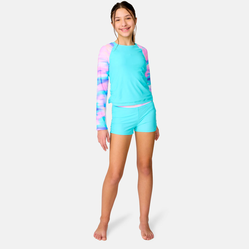 A smiling girl wears the Clearance Girls' Dreamy Drift Rash Guard Swim Set, featuring a turquoise UPF 50+ top with pink and blue tie-dye sleeves and matching quick-dry boy shorts, standing barefoot against a white background.