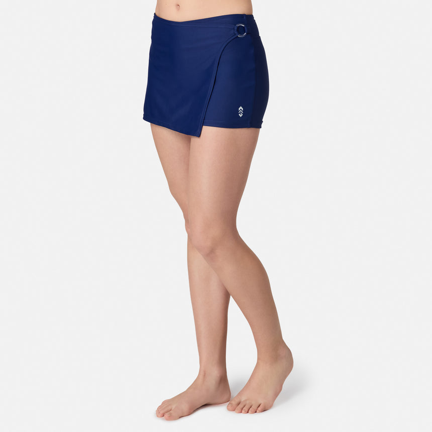 A person stands barefoot in Free Country's Women's Fully Lined Swim Skort, featuring a navy blue color, wrap design, and side ring detail, set against a plain white background.